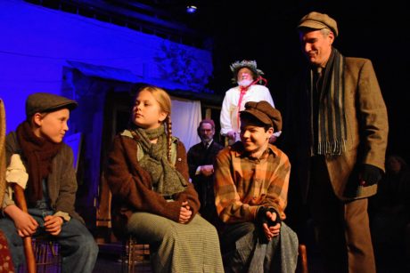 L-R: Caden Mitchell (Tiny Tim), Erin Casey (Belinda Cratchit), Brendan Dure (Peter Cratchit) and Daniel Lavanga (Bob Cratchit) as David Whitehead (Scrooge) and Ward Kay (Ghost of Christmas Present) look on in the Providence Players' production of A Christmas Carol. Photo by Chip Gertzog.